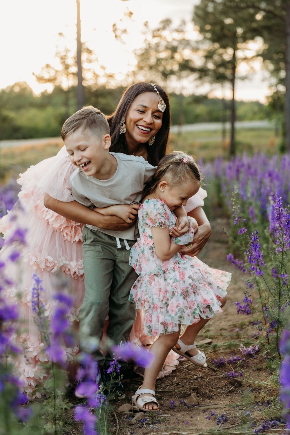 A joyful mother laughs with her two young children in a purple flower field at sunset.