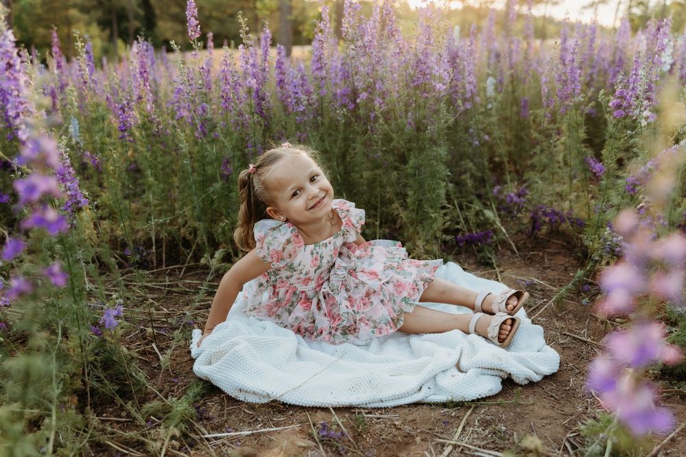 Young girl in floral dress sitting on white blanket surrounded by purple wildflowers in a garden field.