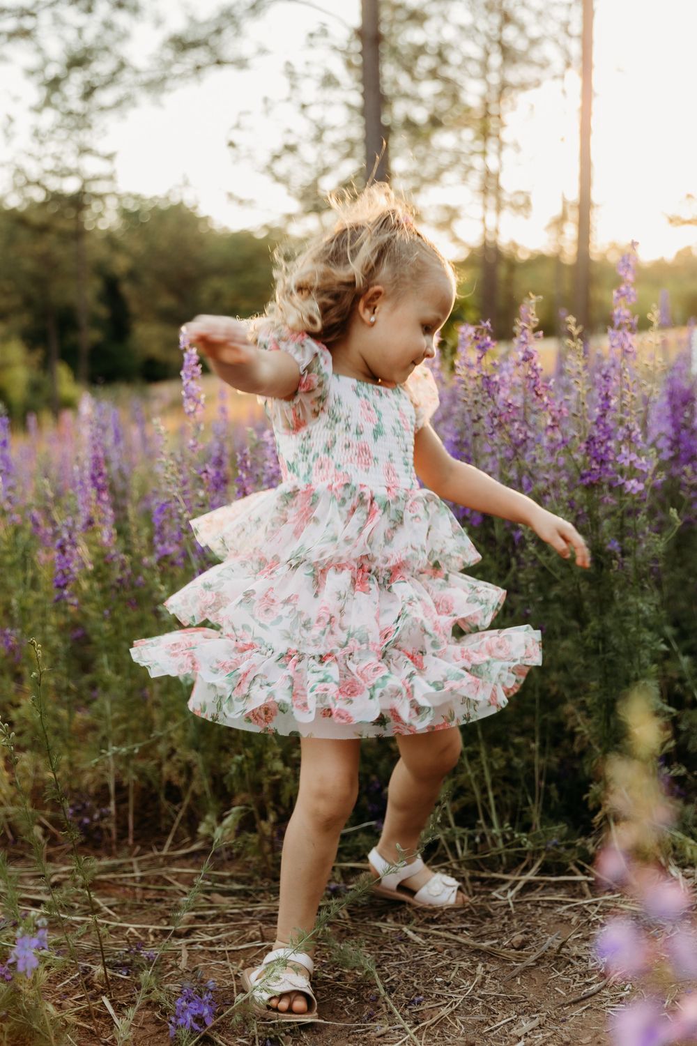 A toddler girl twirls in a floral ruffle dress among purple wildflowers at golden hour.