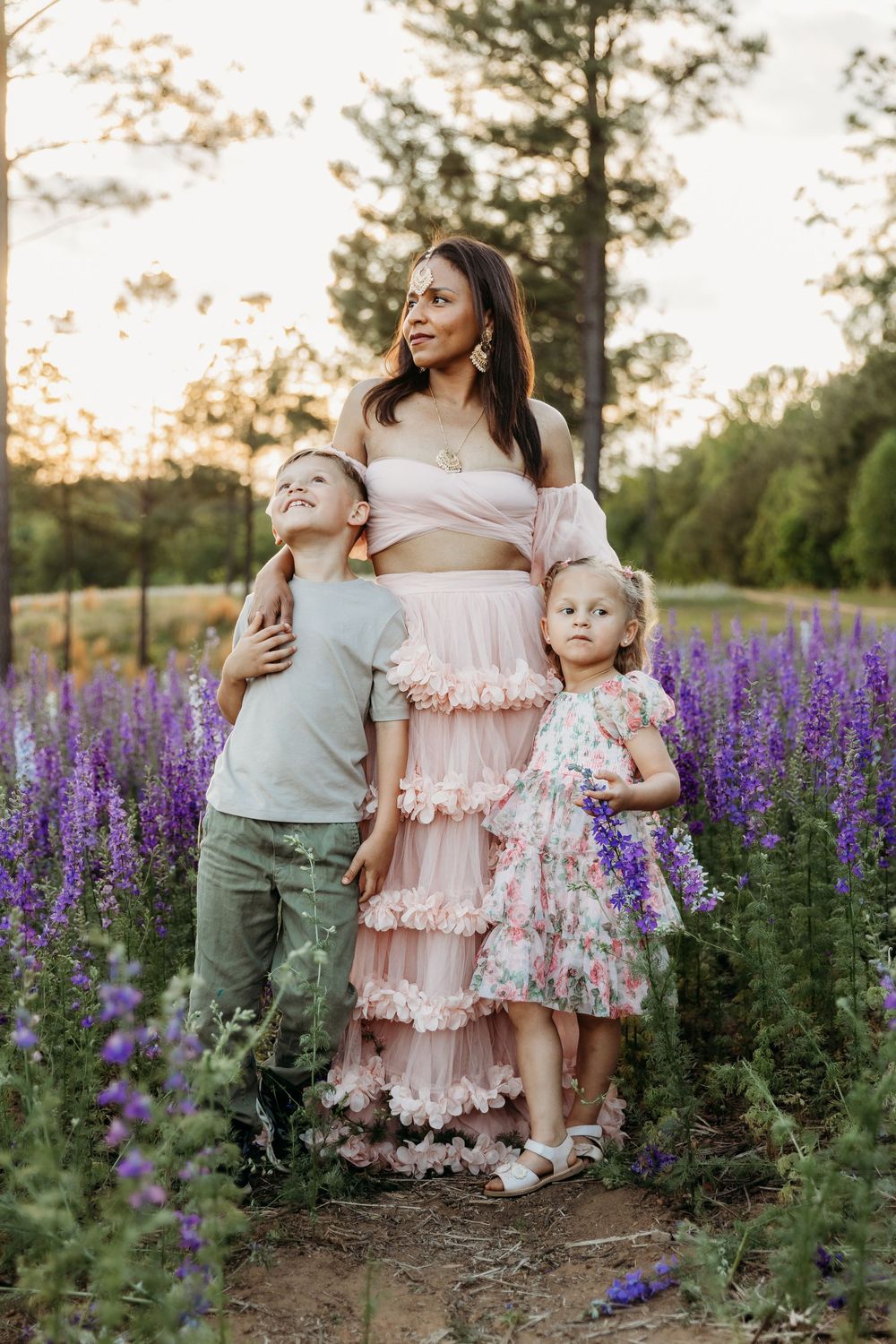 A mother in a pink ruffled dress poses with her two young children in a purple wildflower field at golden hour.