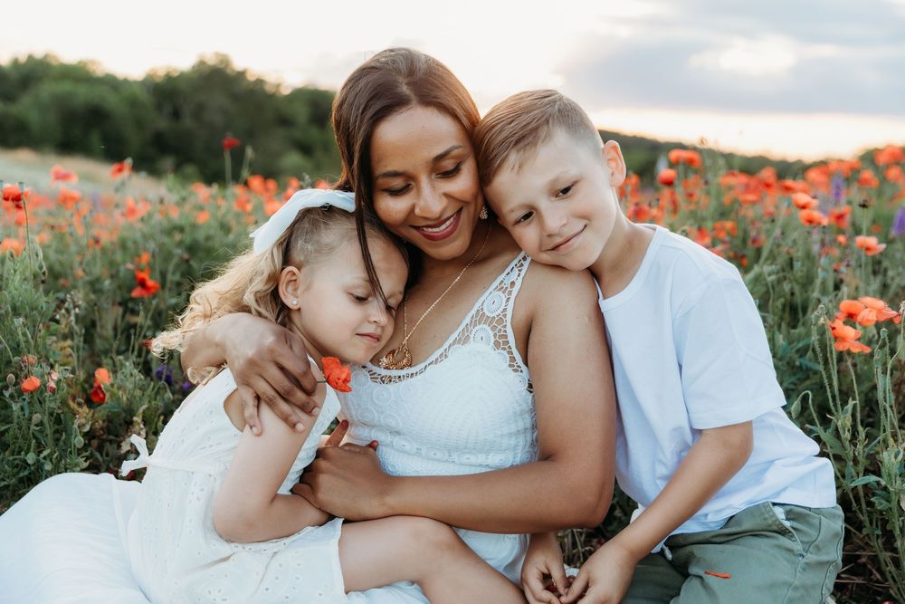 A mother embraces her two children in a blooming poppy field at sunset, all wearing white outfits.