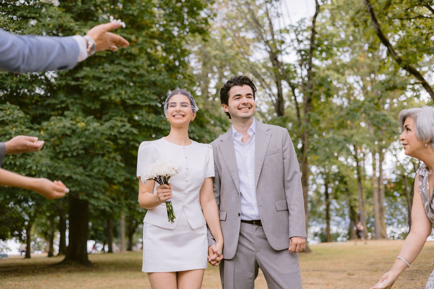 Newlywed couple holding hands in a park, bride in white mini dress with bouquet, groom in gray suit, guests reaching out.
