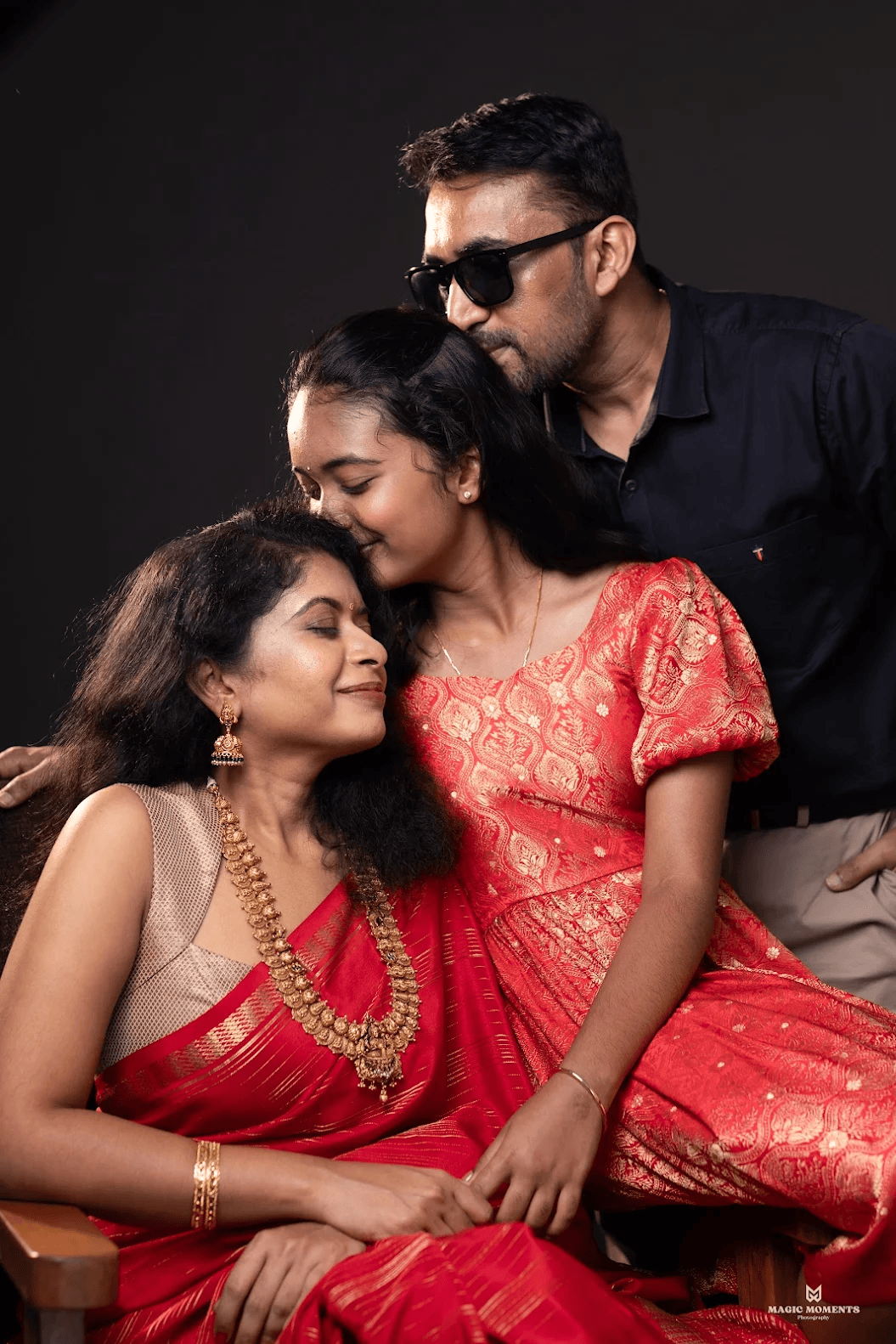 A family of three in traditional Indian attire poses warmly in a studio, wearing red silk garments and gold jewelry.