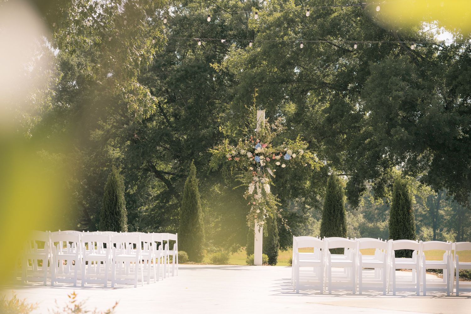Outdoor wedding ceremony setup with white chairs, floral arch, and lush green trees in a sunlit garden venue.