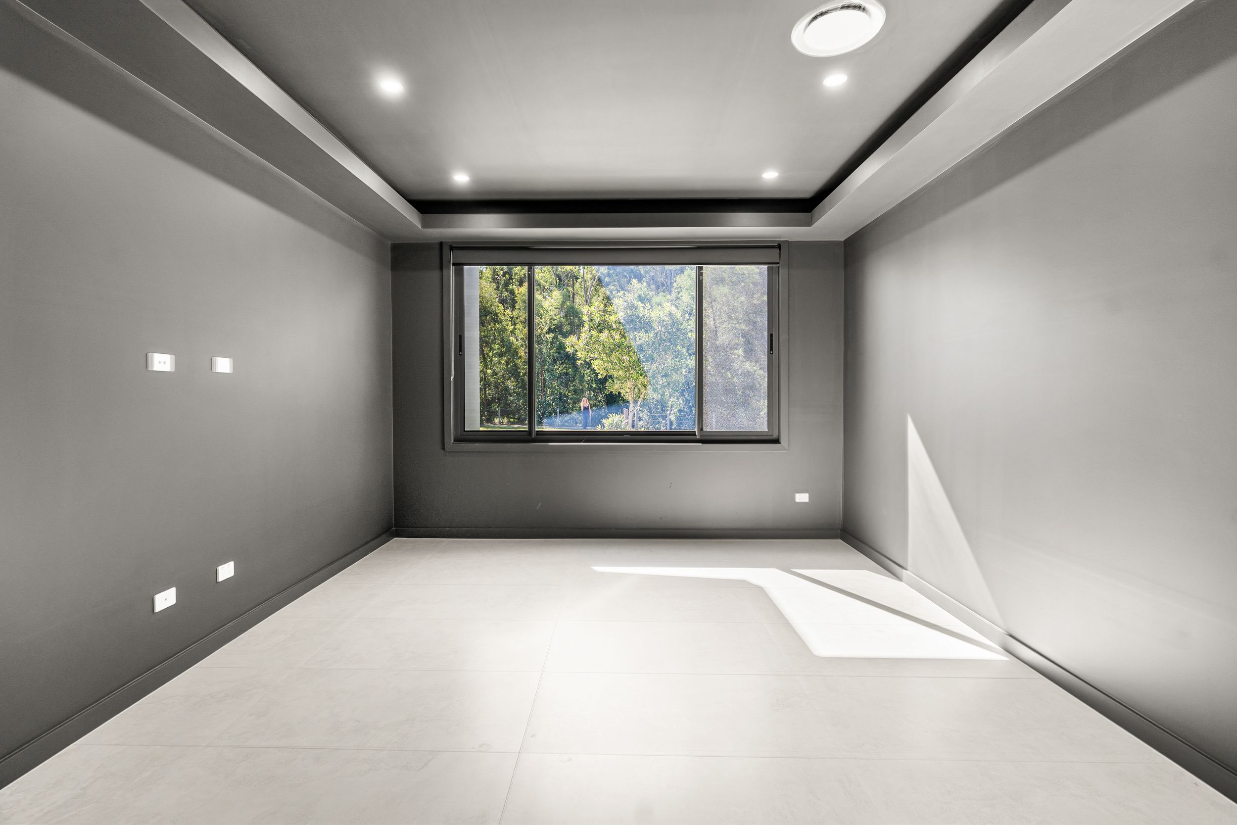 Modern empty room with grey walls, recessed ceiling, large window with garden view, and tiled floor.