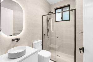 Contemporary ensuite bathroom with round LED mirror, vessel sink, glass shower enclosure, and neutral stone tiles.