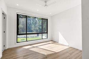 Sunlit empty bedroom with large black-framed picture window offering serene garden views and light timber flooring.