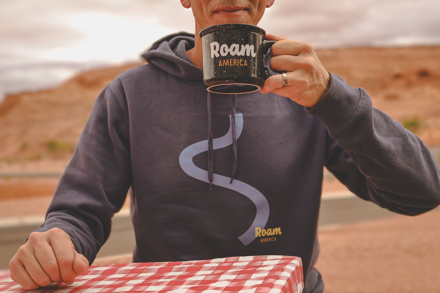 Person in gray Roam America hoodie drinking from a branded mug at a picnic table in the desert.