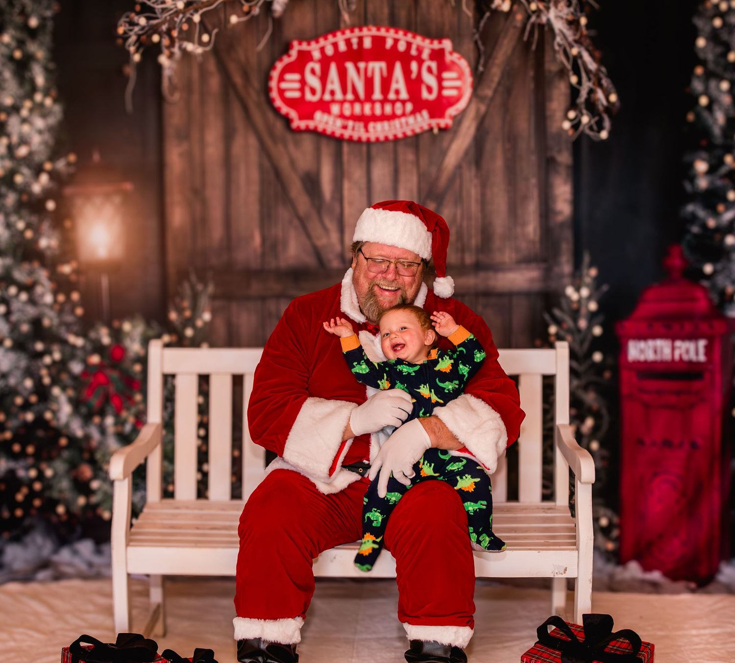 A crying baby in Christmas pajamas sits on Santa Claus's lap in front of a festive Santa's Workshop backdrop.