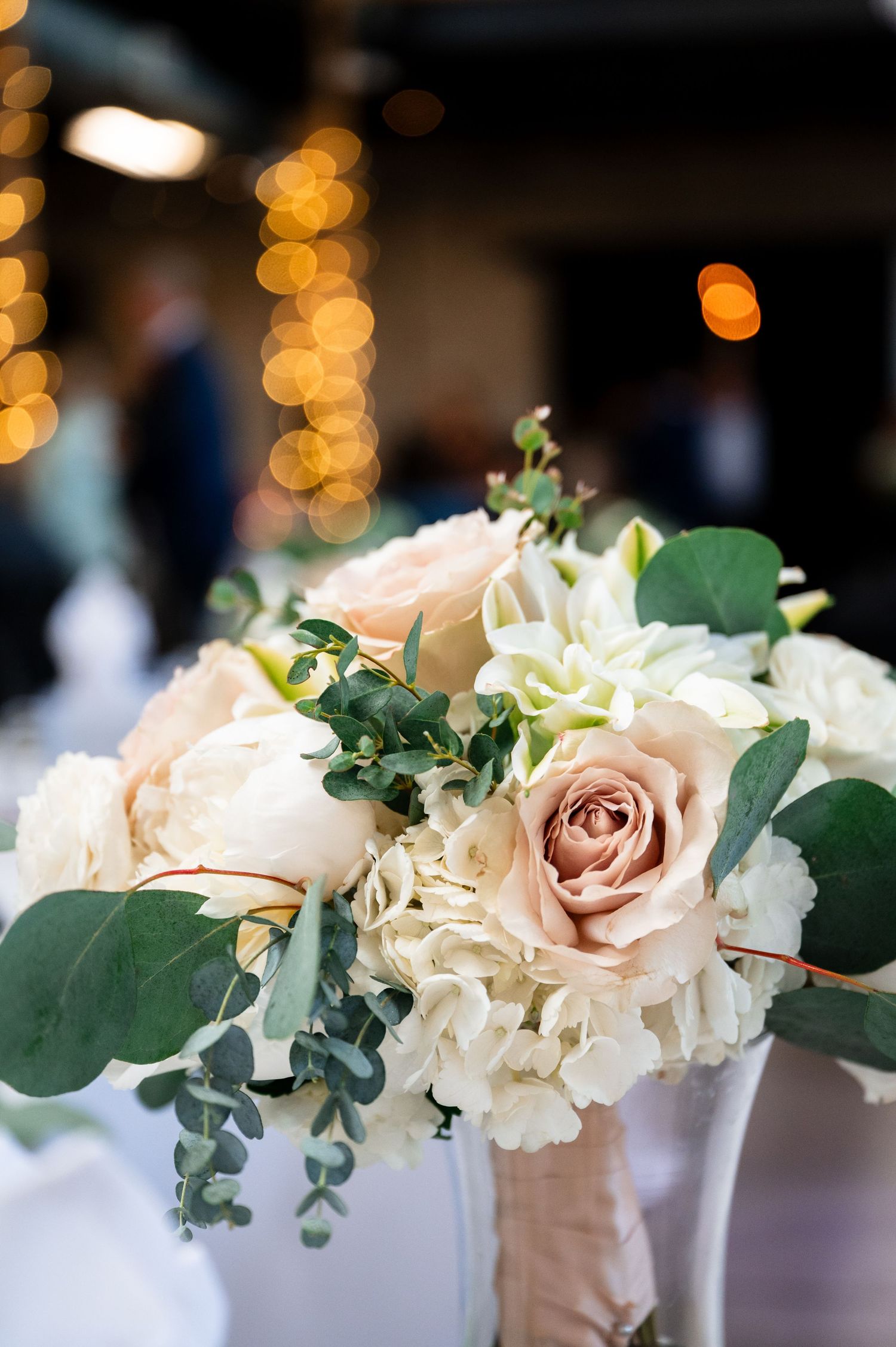 Elegant wedding bouquet with blush roses, white hydrangeas, and eucalyptus greenery, set against a softly lit reception venue.