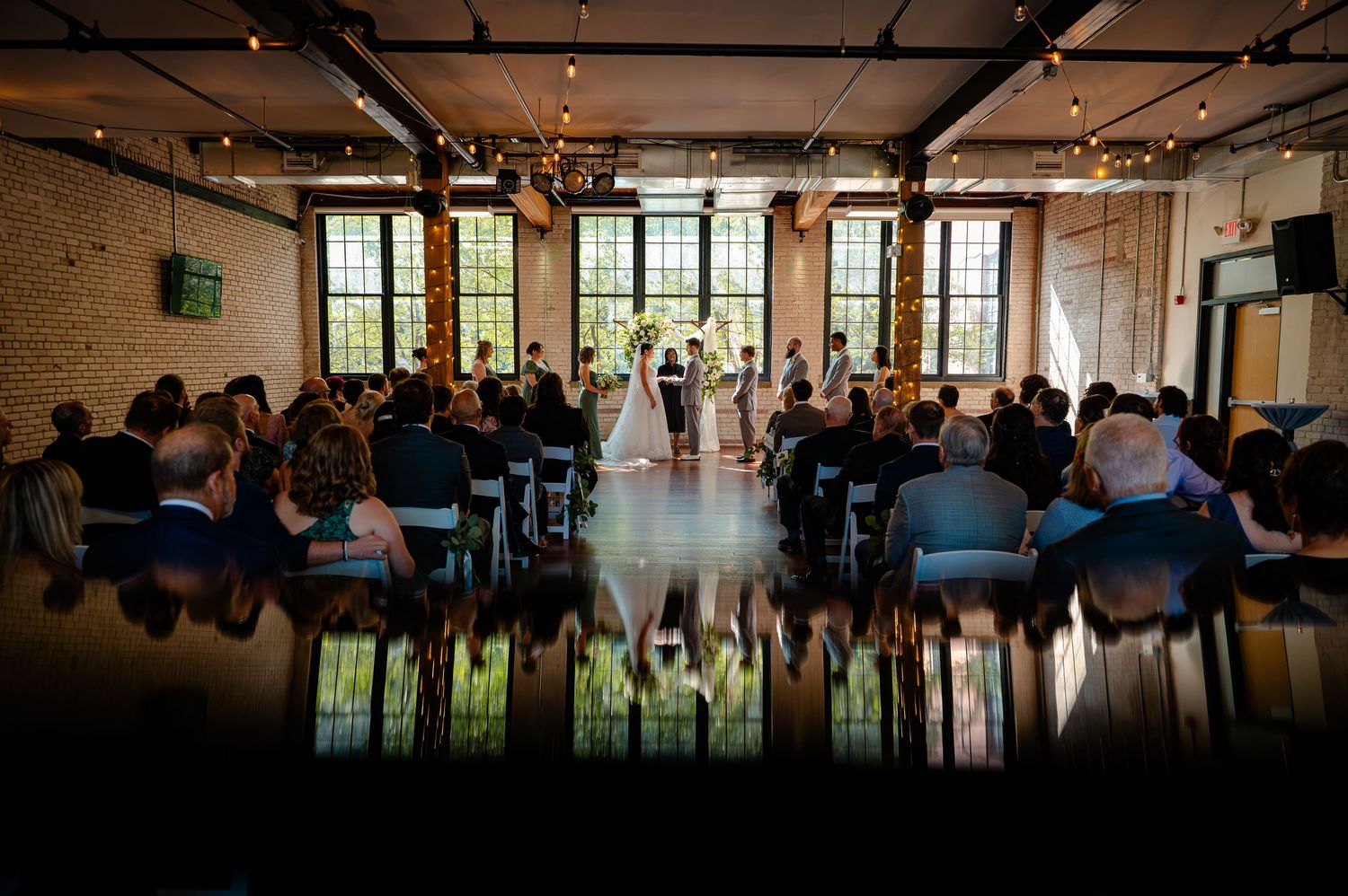 Elegant indoor wedding ceremony in a rustic brick venue with string lights and large windows.