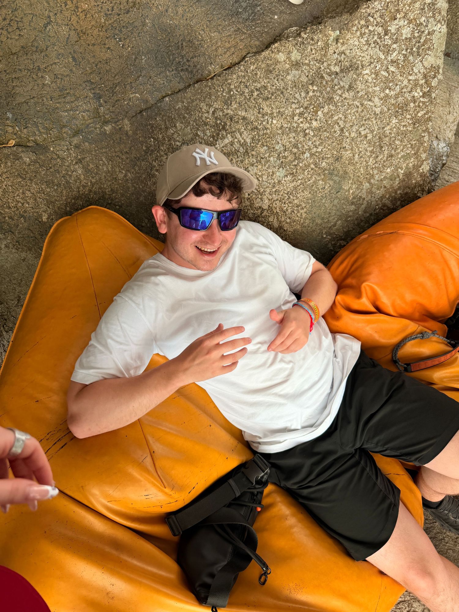 Young man relaxing on orange leather chair wearing NY Yankees cap, blue mirrored sunglasses, white tee, and black shorts.