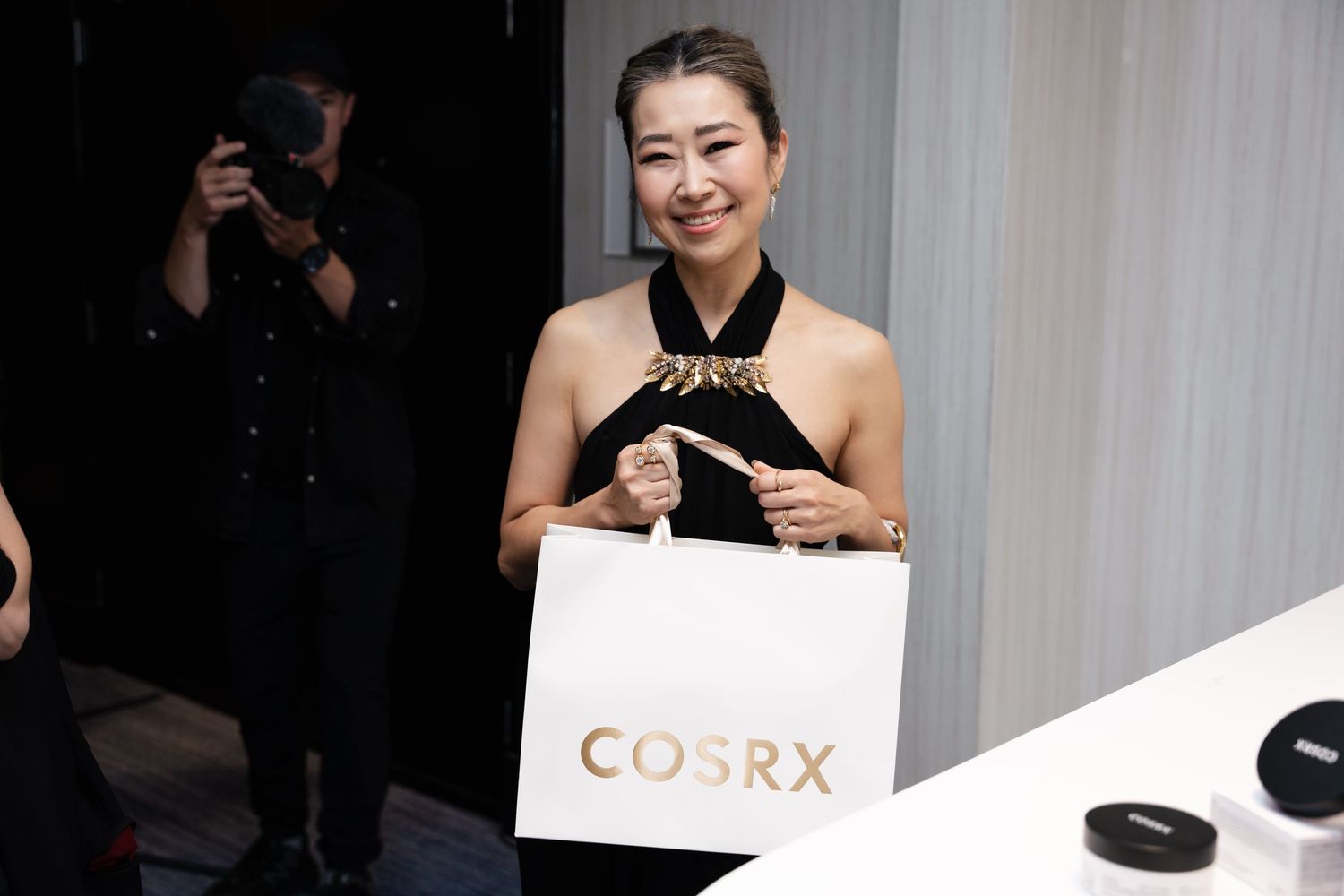Woman smiling and holding a white COSRX shopping bag at a beauty brand event.