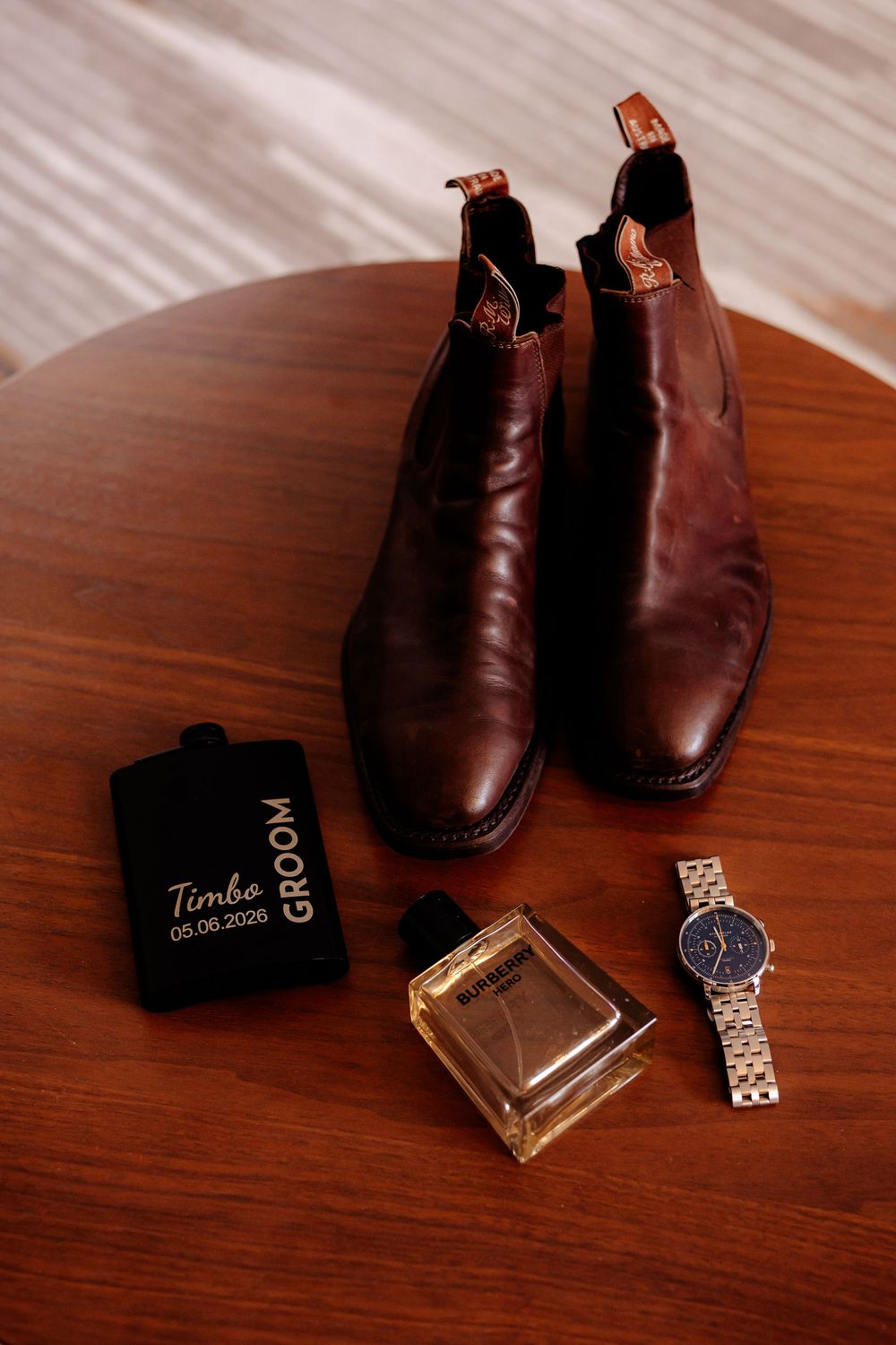 Brown leather Chelsea boots with groom flask, Baldessarini cologne, and silver watch on wooden table for wedding day.