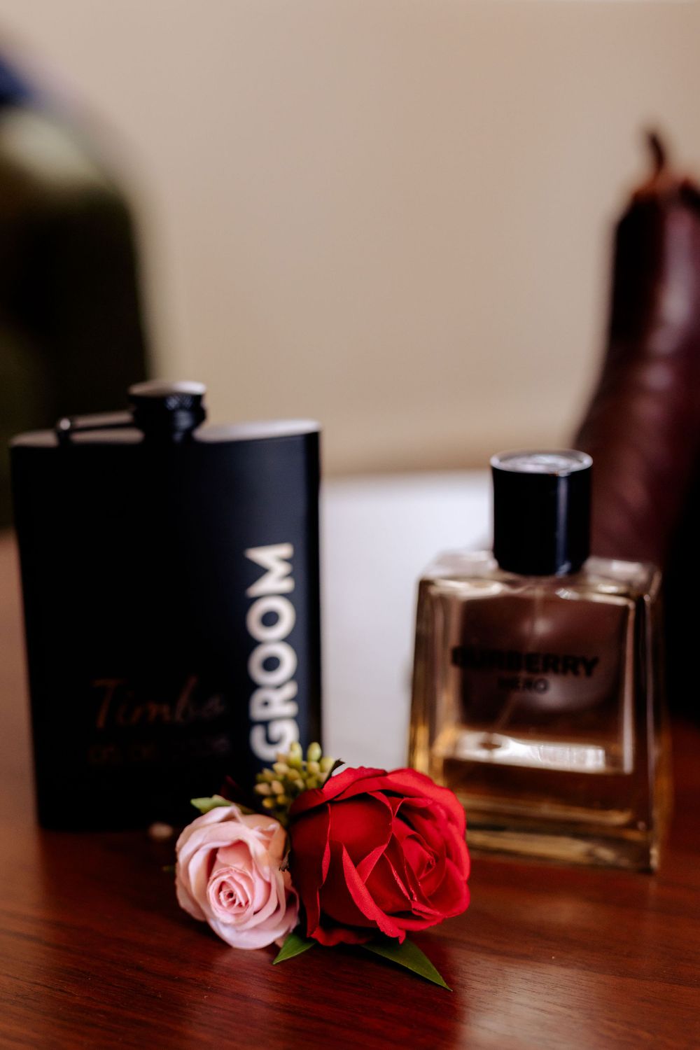 Black groom flask, red and pink roses boutonniere, and Burberry Hero cologne arranged on a wooden table for a wedding.