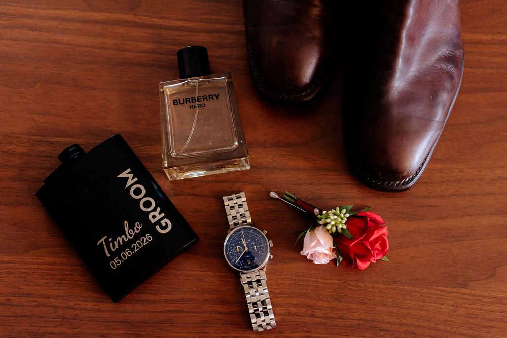Burberry cologne, personalized flask, silver watch, red boutonniere, and brown leather dress shoes on wooden surface.