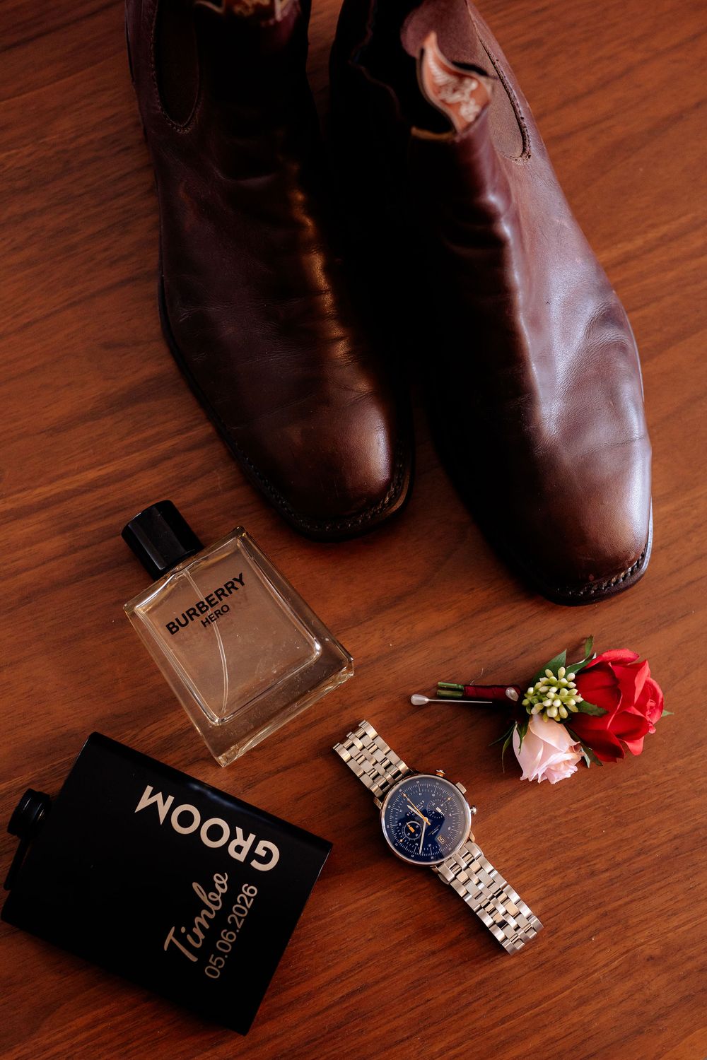 Groom wedding flat lay featuring brown leather boots, Burberry cologne, a silver watch, boutonniere, and personalized black flask.