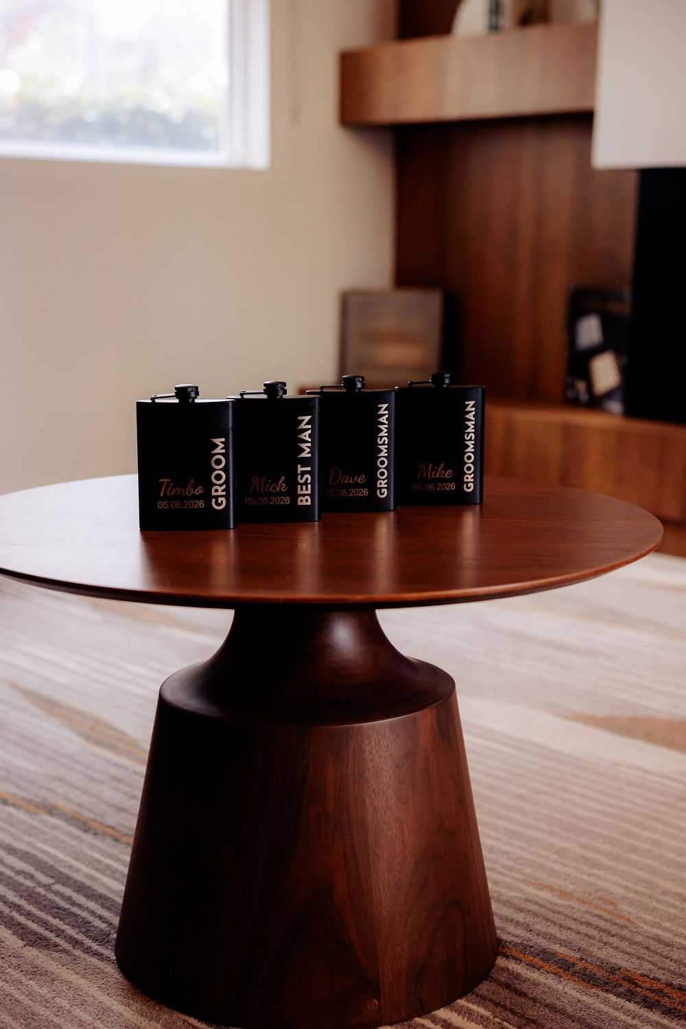 Three personalized black flasks labeled Groom, Best Man, and Groomsman displayed on a round wooden pedestal table.