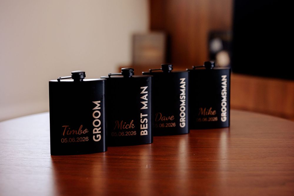Four black engraved flask groomsmen gifts labeled Groom, Best Man, and Groomsmen displayed on a wooden table.