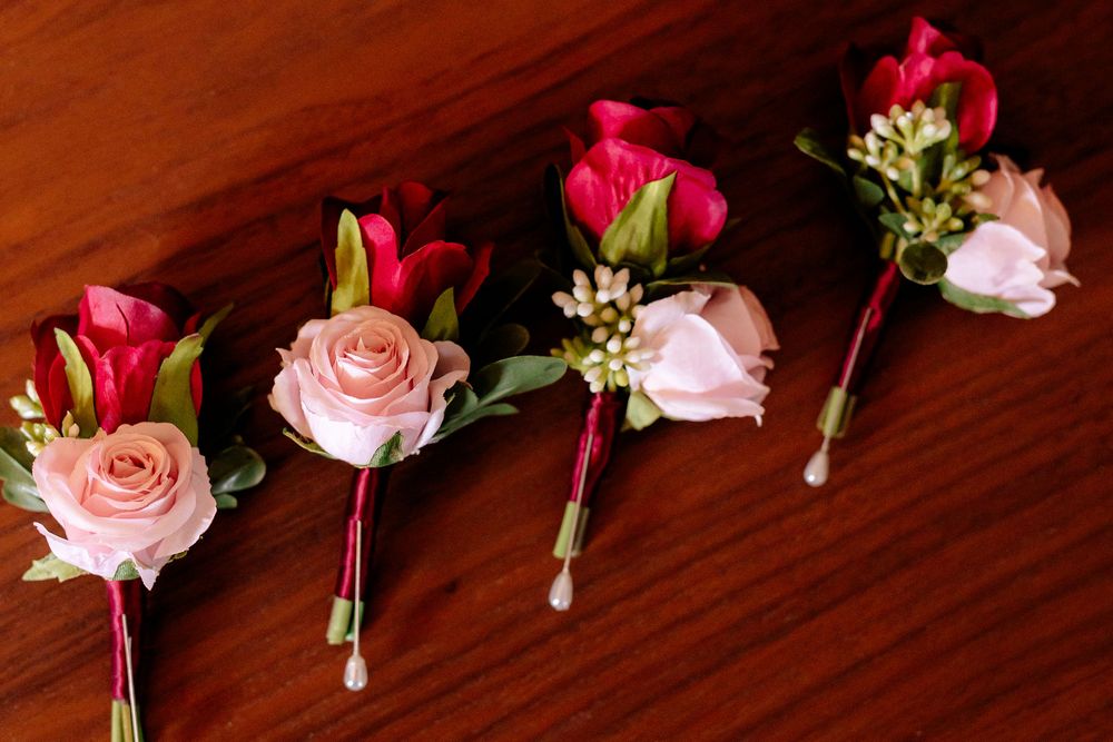 Four elegant pink and red rose boutonnieres with greenery arranged in a row on a wooden surface.