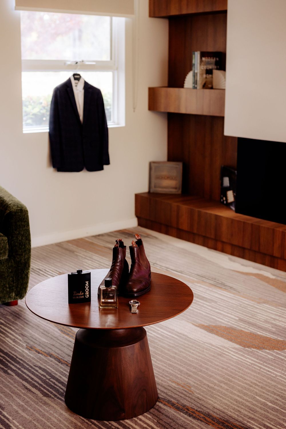 A dark suit jacket hangs by a window beside a wooden coffee table with brown leather boots and cologne in a modern room.