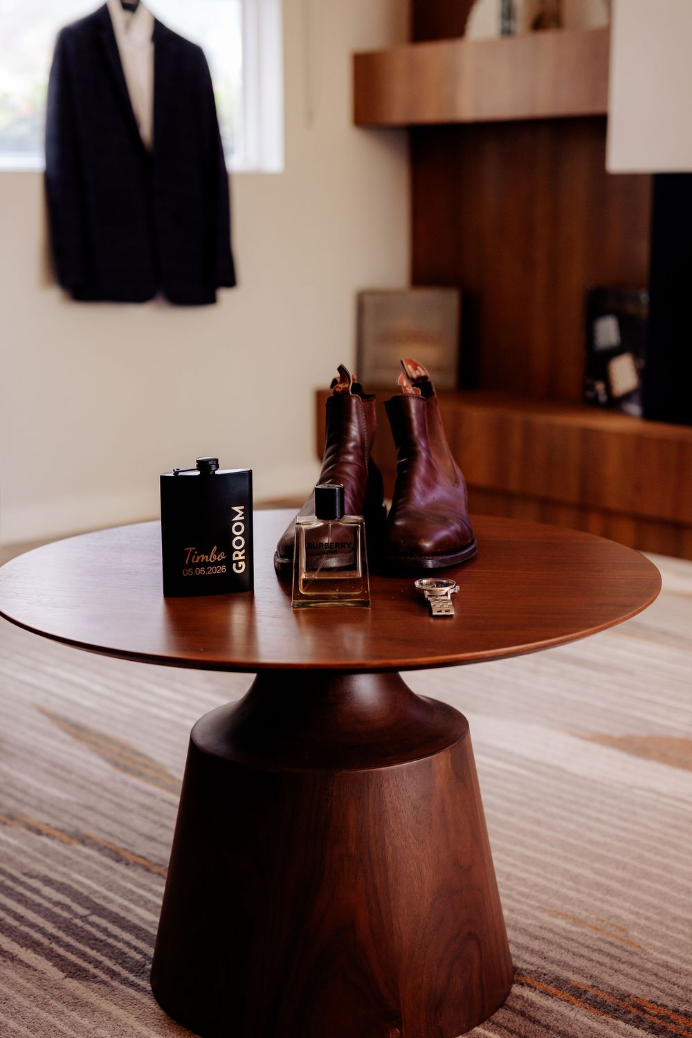 Groom's wedding accessories on walnut pedestal table, including flask, cologne, ring, and brown leather boots.