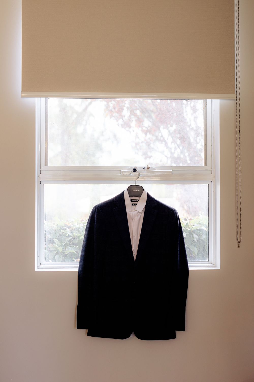 Dark navy suit jacket and white dress shirt hanging on a hanger in front of a bright window with garden views.