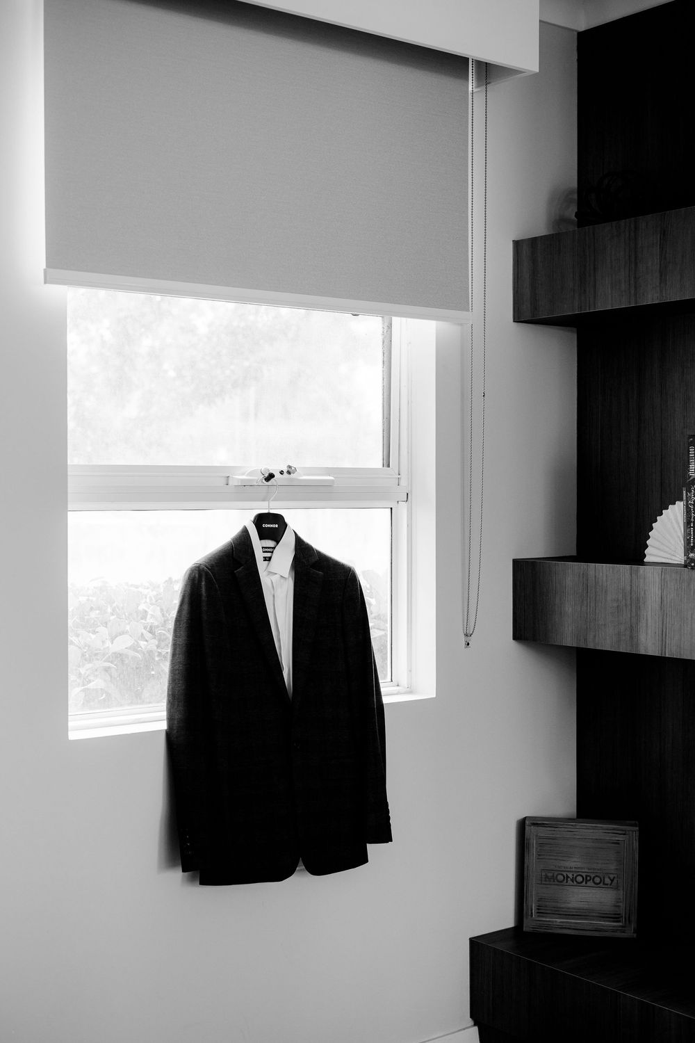 Black and white photo of a suit jacket hanging on a window hanger beside dark wooden shelves in a modern room.