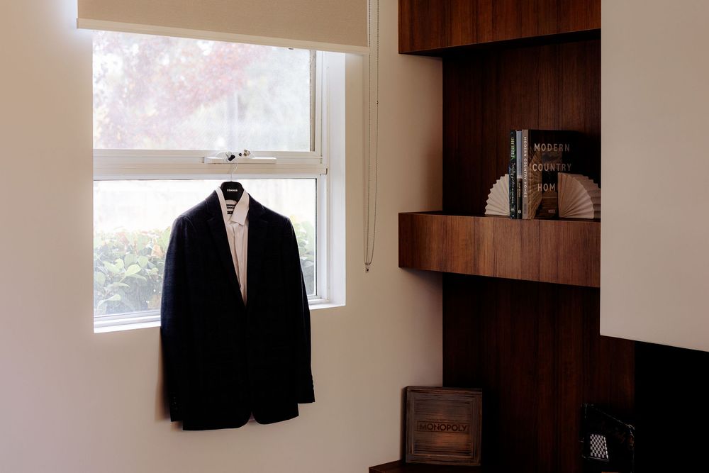 A dark suit jacket on a mannequin hangs near a bright window beside a wooden built-in shelving unit with books.
