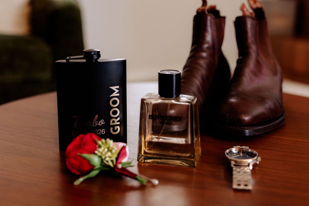 Groom accessories flat lay featuring cologne, flask, boutonniere, watch, and brown Chelsea boots on a wooden table.