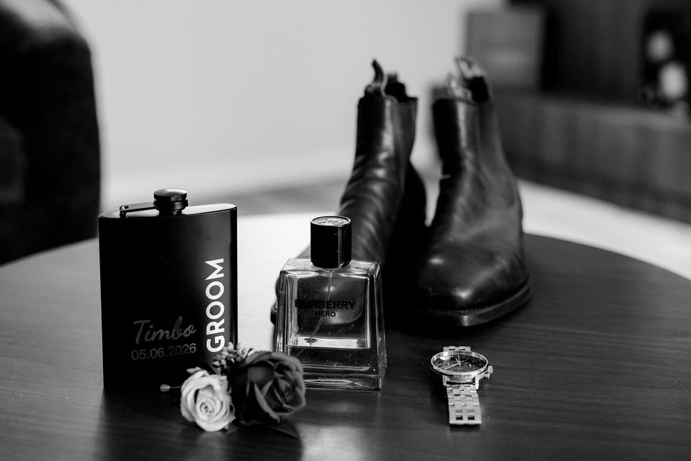 Black and white flat lay of groom wedding accessories including flask, cologne, watch, cufflinks, and leather Chelsea boots.
