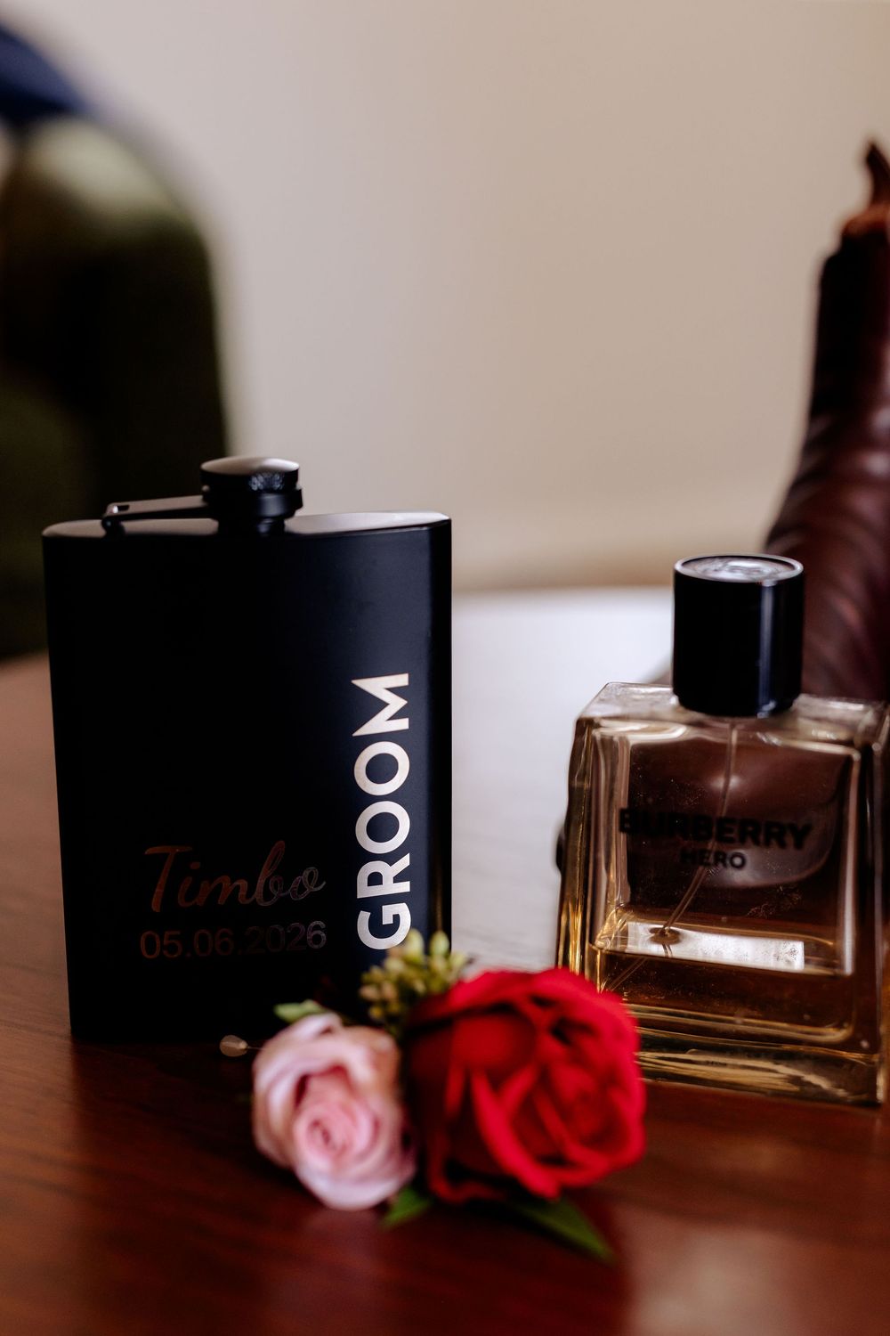 Black groom flask engraved with Timbo date beside Burberry Hero cologne and red roses on wooden table.