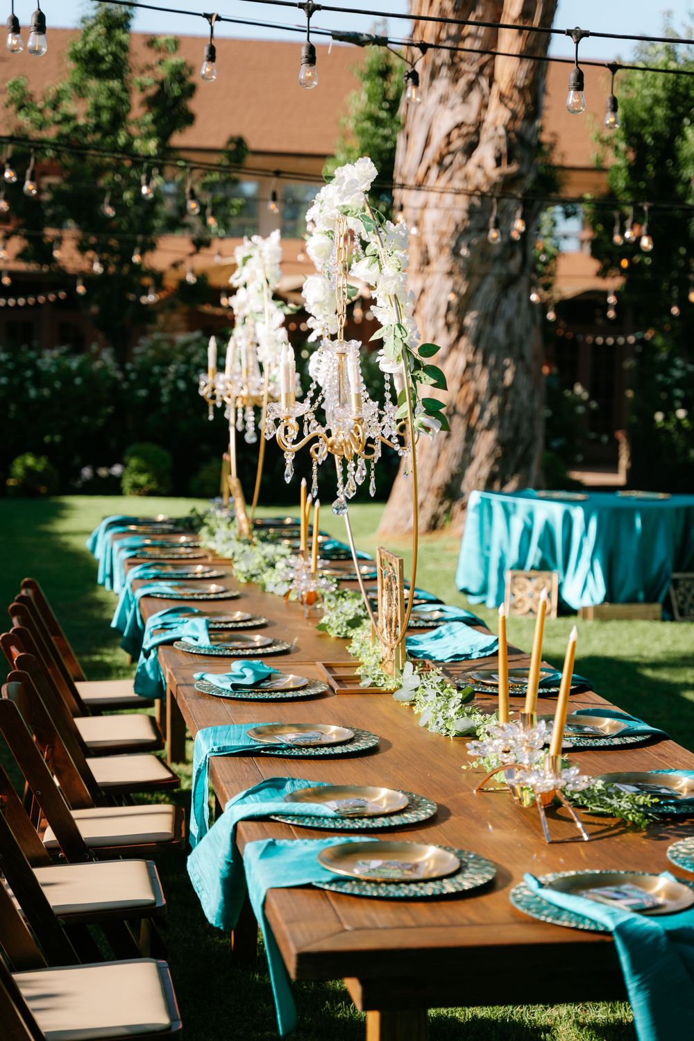 Elegant outdoor wedding reception table with teal napkins, gold candles, crystal chandeliers, and floral centerpieces under string lights.