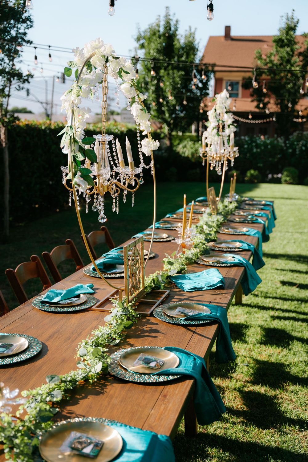 Elegant outdoor wedding reception table with teal napkins, gold chargers, crystal chandeliers, and greenery garland on a wooden farm table.