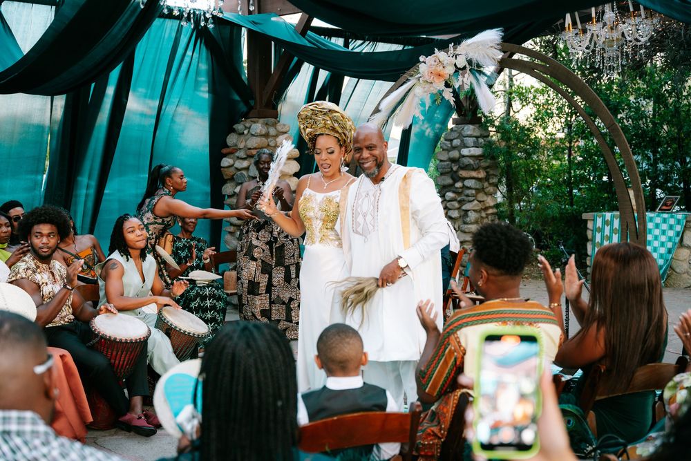 Couple in traditional African wedding attire celebrate ceremony with guests playing drums outdoors.