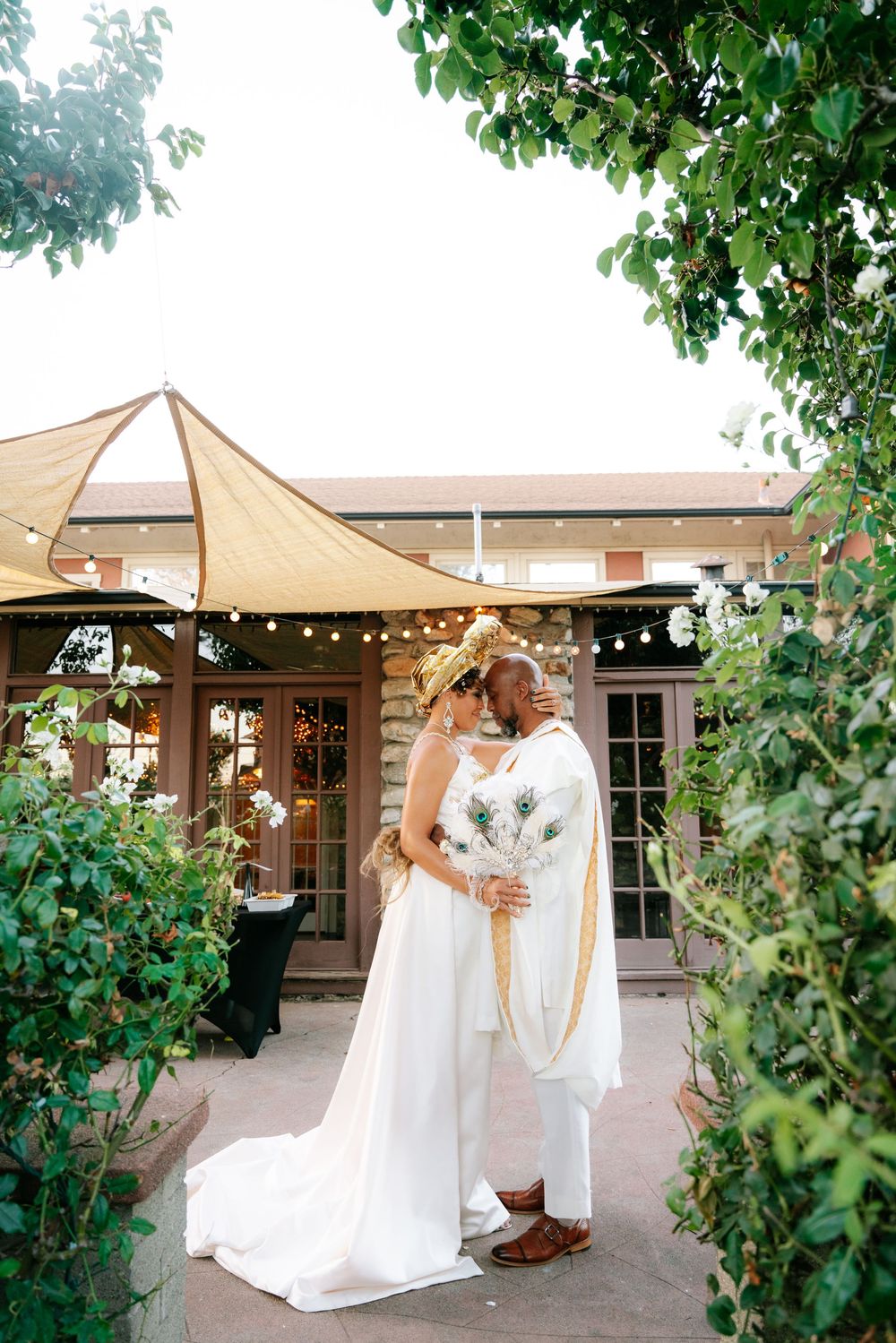 Bride and groom share a romantic moment in a lush garden courtyard, bride holding a peacock feather bouquet.