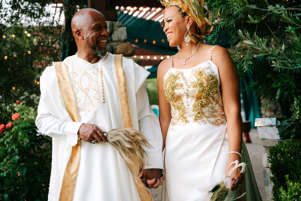 Couple in elegant white and gold wedding attire holding hands outdoors, smiling joyfully at a garden ceremony.