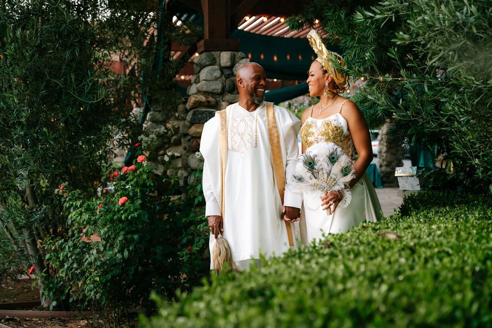 Couple in elegant white and gold traditional African attire walking through a lush green garden setting.