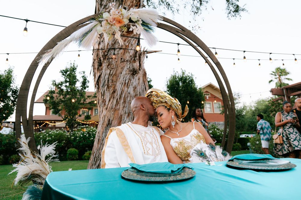 Couple in traditional attire sharing a romantic moment at a turquoise table under a circular arch outdoors.