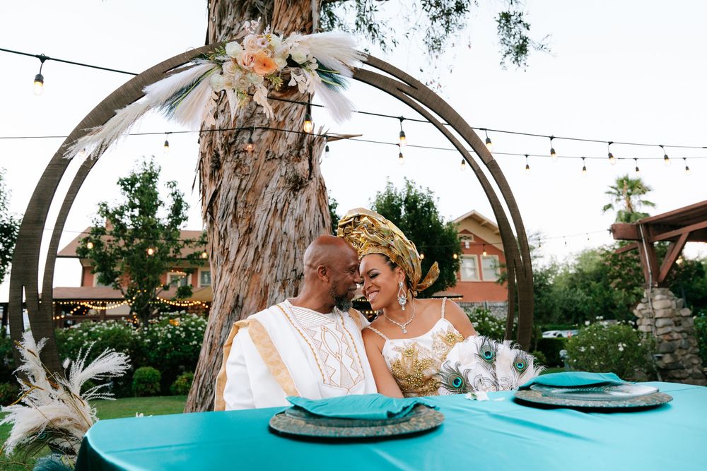African couple in traditional attire shares romantic moment under floral arch at turquoise wedding table outdoors.