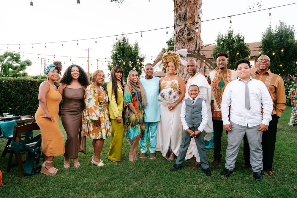 Diverse group of elegantly dressed wedding guests posing together outdoors under string lights on a green lawn.