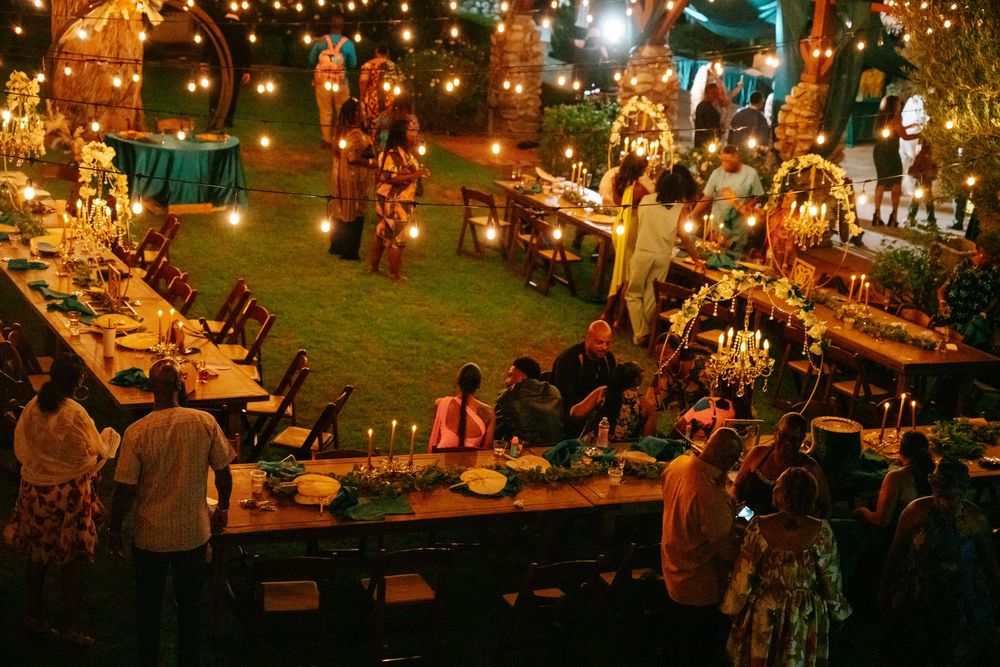 Elegant outdoor evening wedding reception with string lights, long dining tables, candles, and guests mingling on lush green lawn.