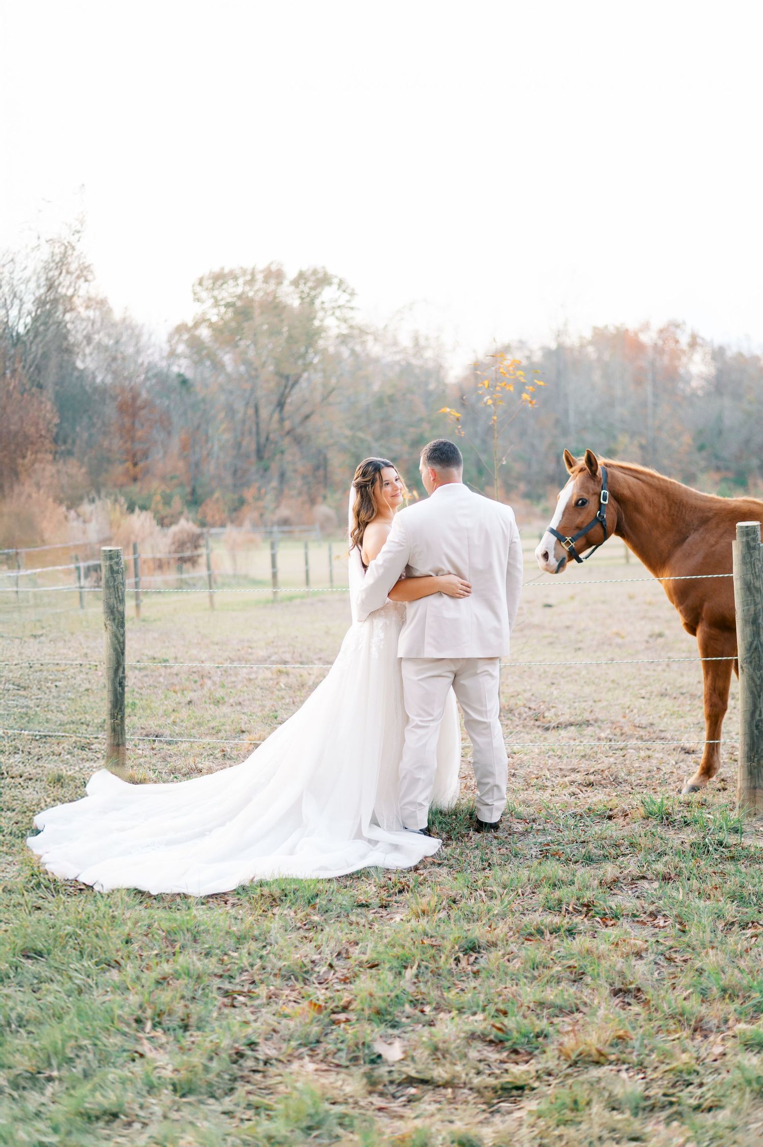 Groom in sage suit kisses bride's temple as she smiles holding colorful pink and white floral bouquet outdoors.