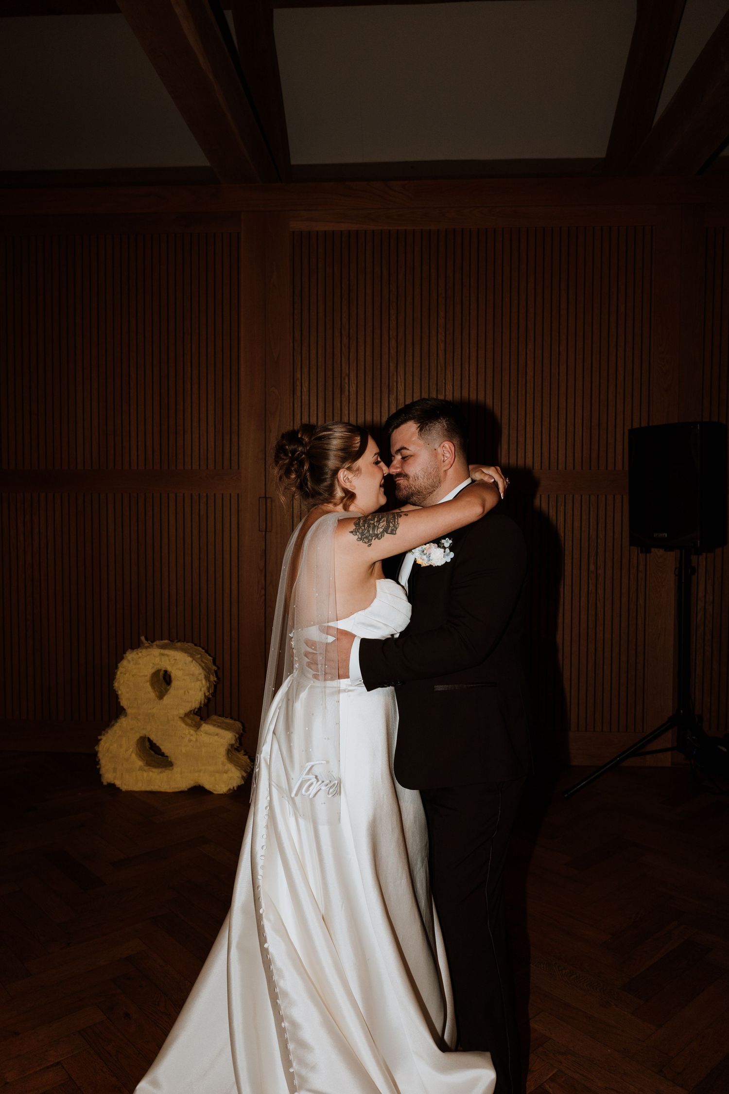 Bride and groom share their first dance, embracing tenderly in a warmly lit reception hall with a glowing ampersand sign.