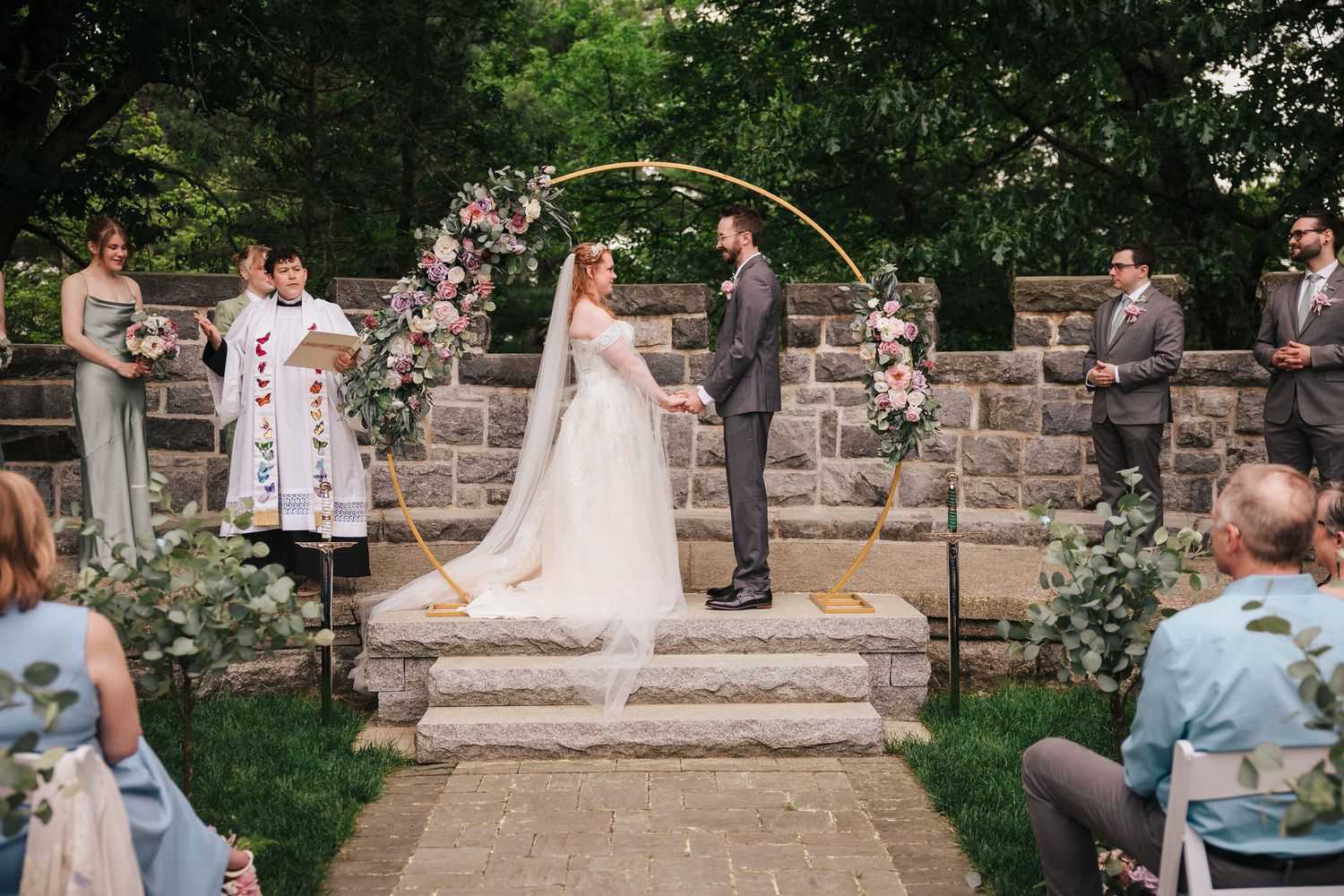 Bride and groom holding hands at the altar under a floral arch, full wedding party gathered at Searles Castle.