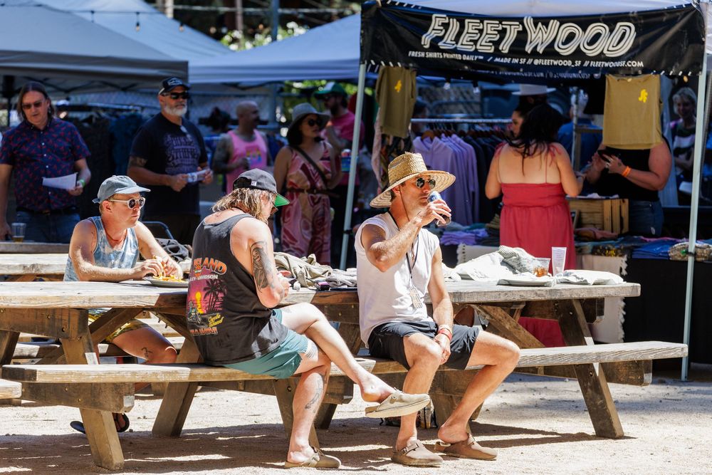 People relaxing at picnic tables outdoors near a Fleetwood vendor tent at a sunny festival event.