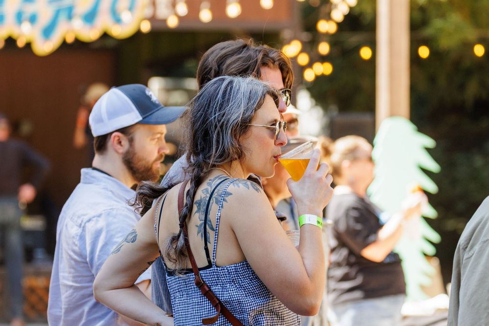 Woman sipping craft beer at outdoor festival with friends socializing in background under string lights.