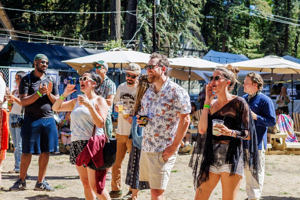 Diverse group of happy people enjoying an outdoor festival on a sunny day with market stalls and trees in background.