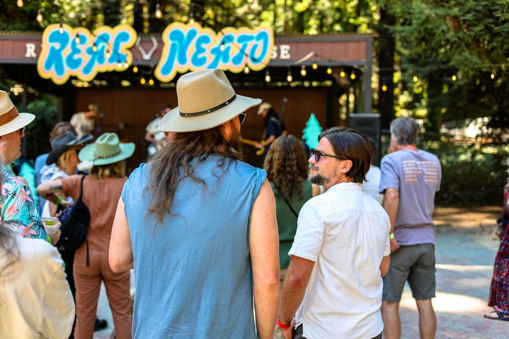 Crowd of festival-goers gathered outdoors near a colorful Real Neato stage sign surrounded by tall trees.