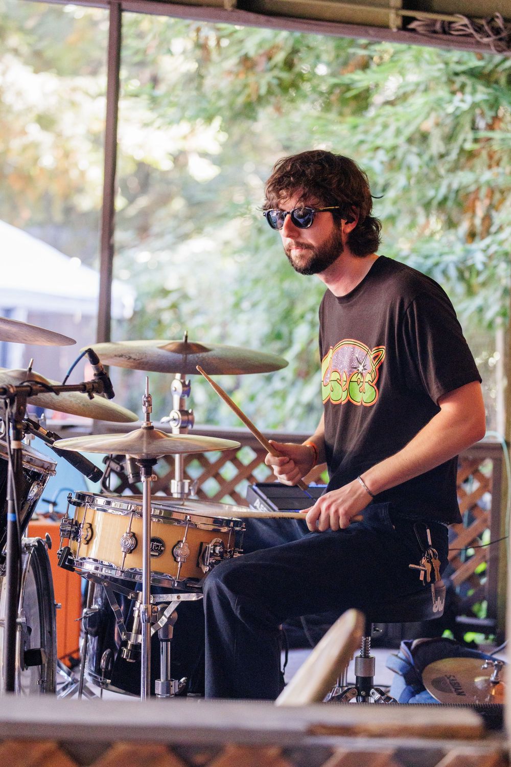 Young bearded drummer with sunglasses playing drum kit on outdoor stage during live music performance.